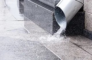 Rainwater pouring out of a metal downspout onto pavement next to a building wall